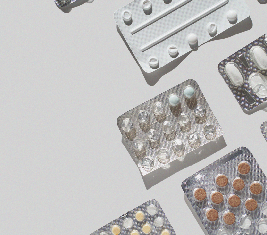 Flat lay of assorted blister packs containing various pills and tablets arranged diagonally on a light gray background, casting soft shadows in natural light.