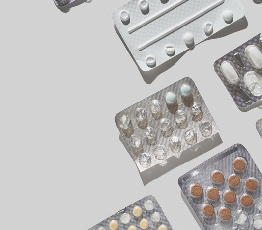 Flat lay of assorted blister packs containing various pills and tablets arranged diagonally on a light gray background, casting soft shadows in natural light.