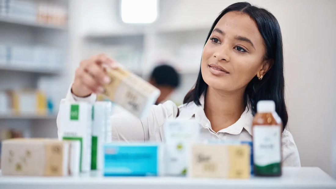 A person in a pharmacy wearing a white shirt reaches for a medicine box from a shelf, with various medication boxes and a bottle of liquid medicine visible in the foreground. The background is softly blurred, showing additional pharmacy shelves.