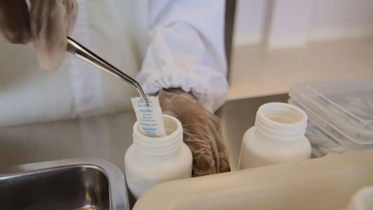 A gloved worker uses metal tweezers to place a moisture-absorbing desiccant packet into a white plastic supplement bottle at a packaging station. An open empty bottle and plastic containers are visible nearby. 