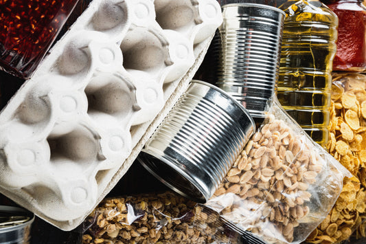 Assortment of packaged pantry foods including canned goods, peanuts, cereal flakes, granola, cooking oil, and an empty egg carton arranged closely together.