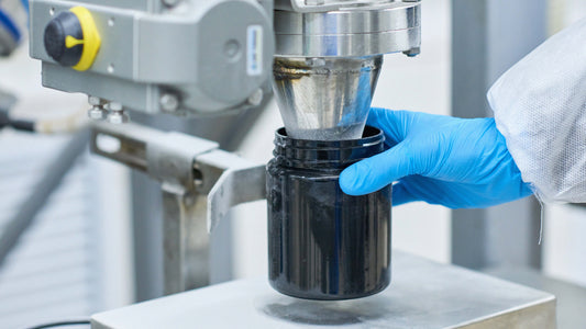 Close-up of a pharmaceutical manufacturing process where a worker wearing blue gloves holds a black container beneath a stainless-steel dispensing machine filling it with powdered material in an industrial lab or production facility.