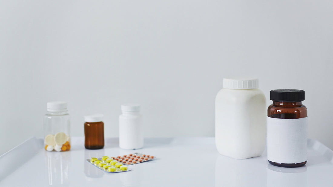 Assorted pharmaceutical packaging on a white surface, including clear and amber pill bottles, white plastic containers, and blister packs with tablets. Minimalist clinical setting with ample empty space in the background.