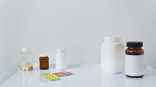 Assorted pharmaceutical packaging on a white surface, including clear and amber pill bottles, white plastic containers, and blister packs with tablets. Minimalist clinical setting with ample empty space in the background.