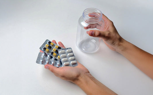 Hands holding multiple blister packs of assorted capsules and tablets next to an empty clear supplement bottle on a white surface.