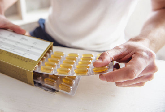 Close-up of a person removing a blister pack of golden softgel capsules from a cardboard supplement box on a white table.