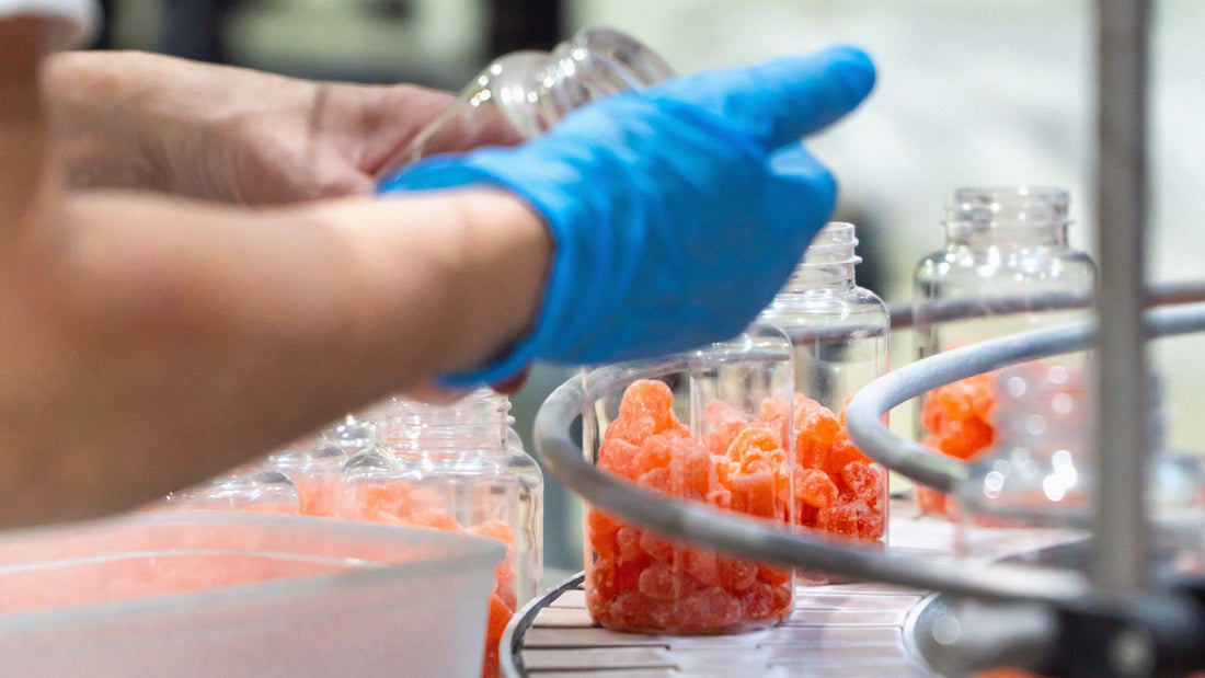 Worker wearing blue gloves fills clear plastic supplement bottles with orange gummy supplements on an automated conveyor belt in a manufacturing facility. Multiple open bottles move through the production line under bright industrial lighting.