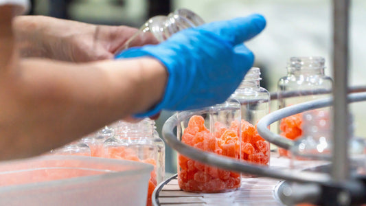Worker wearing blue gloves fills clear plastic supplement bottles with orange gummy supplements on an automated conveyor belt in a manufacturing facility. Multiple open bottles move through the production line under bright industrial lighting.