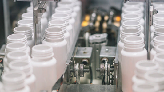 Rows of empty white plastic supplement bottles move through an automated supplement production line with stainless steel machinery and conveyor guides in a manufacturing facility.
