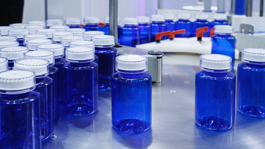 Close-up of blue plastic supplement bottles with white caps moving along a stainless steel conveyor belt in an automated packaging facility. Rows of empty bottles are aligned on the production line under bright industrial lighting.
